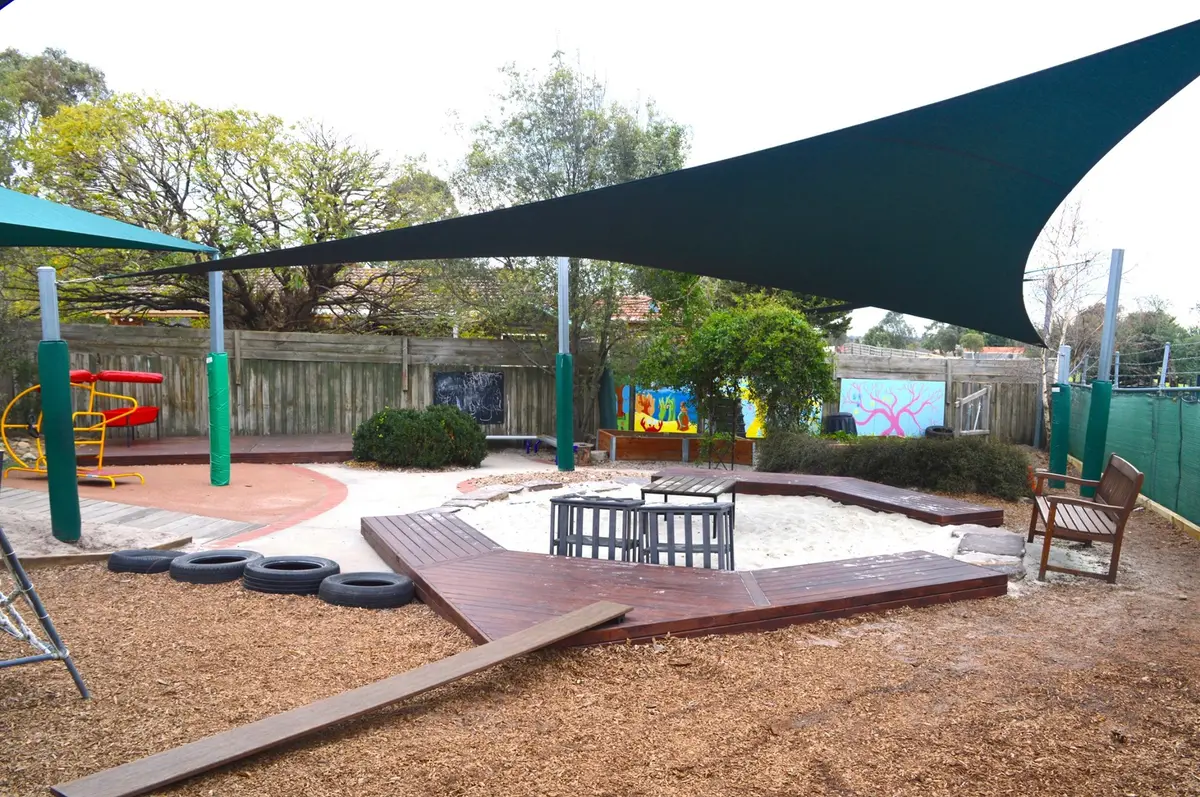 The kinder yard: a sandpit with timber decking around it under a shade sail, a climbing frame and slide on soft fall, painted murals along the back fence and gum trees behind it.